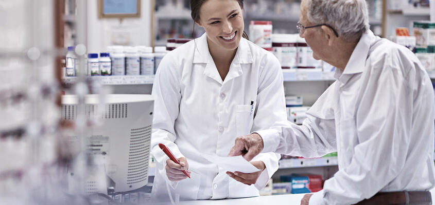 Ein betagter Kunde zeigt einer Apothekerin ein Rezept für Medikamente.